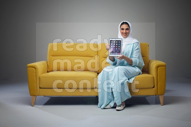 A Saudi woman wearing a light blue Abaya and white head scarf sitting on a yellow sofa and showing her tablet's screen while pointing to it eye level on a grey background