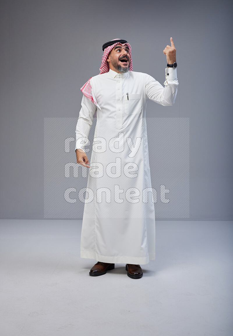 Saudi man Wearing Thob and red white Shomag standing interacting with the camera on Gray background