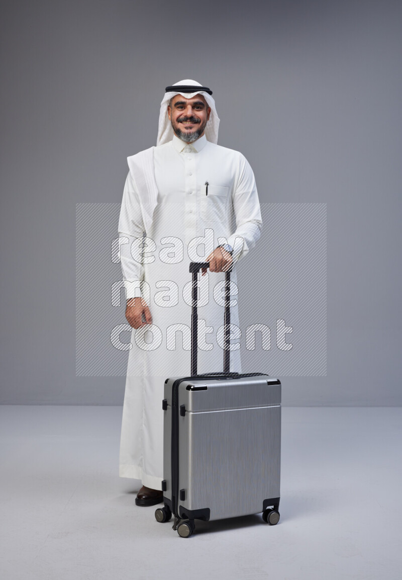 Saudi man wearing Thob and white Shomag standing holding Travel bag on Gray background