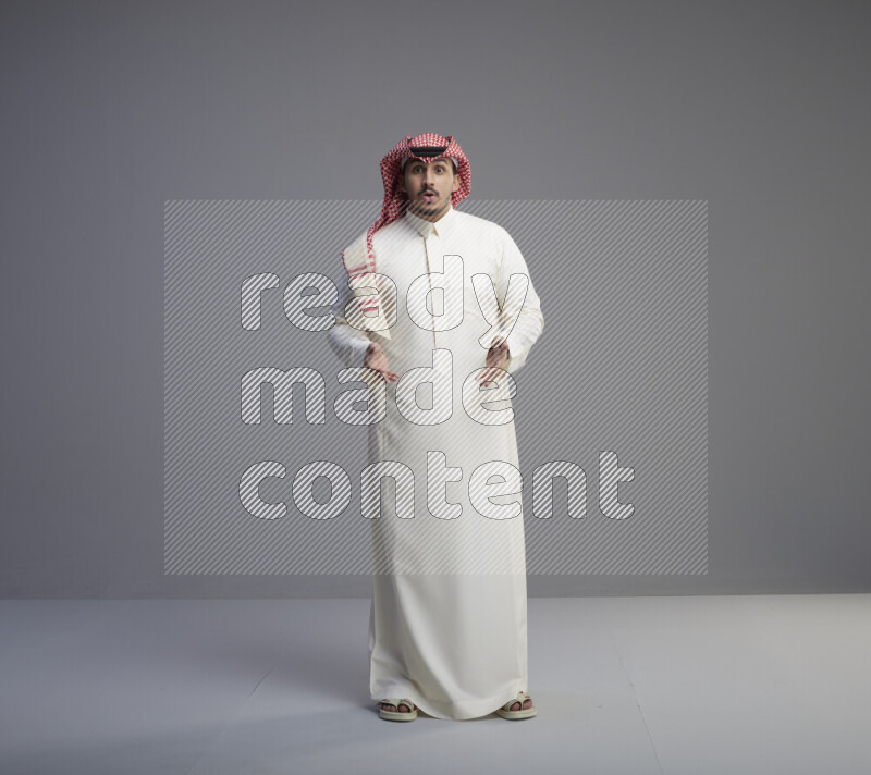 A Saudi man standing wearing thob and red shomag interacting with the camera on gray background