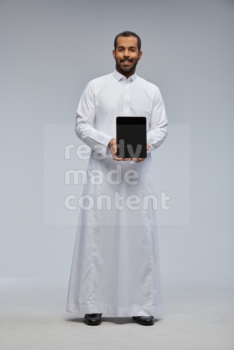 Saudi man wearing Thob standing showing tablet to camera on Gray background