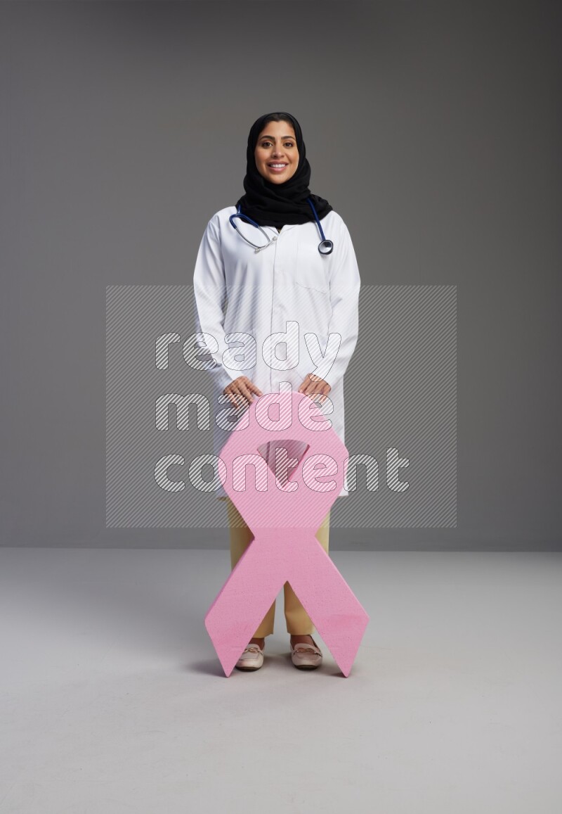 Saudi woman wearing lab coat with stethoscope standing holding awareness ribbon symbol standing on Gray background