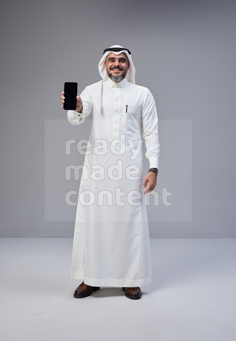 Saudi man Wearing Thob and white Shomag standing showing phone to camera on Gray background