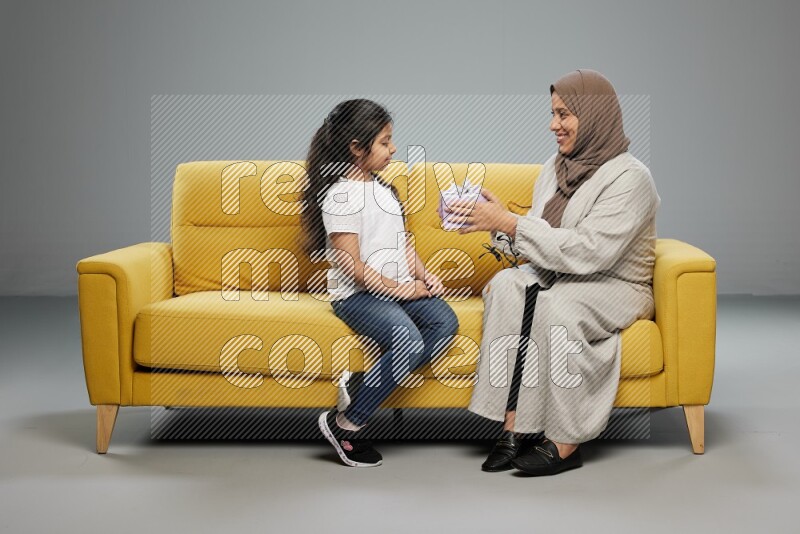 A girl sitting on a yellow sofa and giving a gift to her mother on gray background