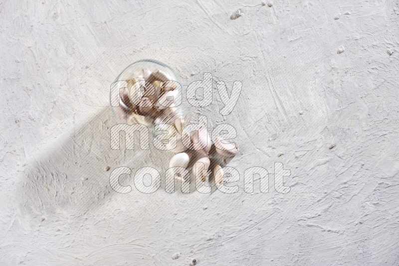 A glass spice jar full of garlic cloves flipped over with the cloves spilling out on a textured white flooring