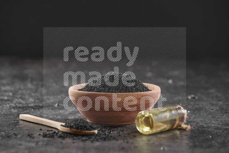 A wooden bowl and spoon full of black seeds and a bottle of black seeds oil on a textured black flooring