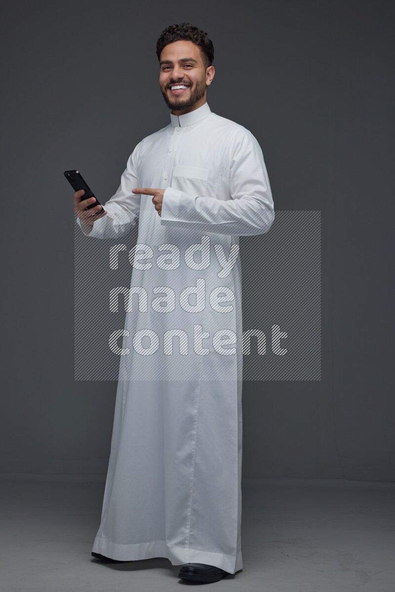 A Saudi man wearing Thobe and holding his phone while pointing to it eye level on a gray background