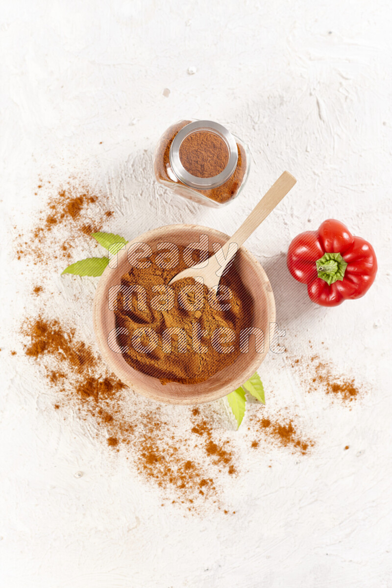 A wooden bowl full of ground paprika powder with a glass jar beside it and red bell pepper on white background