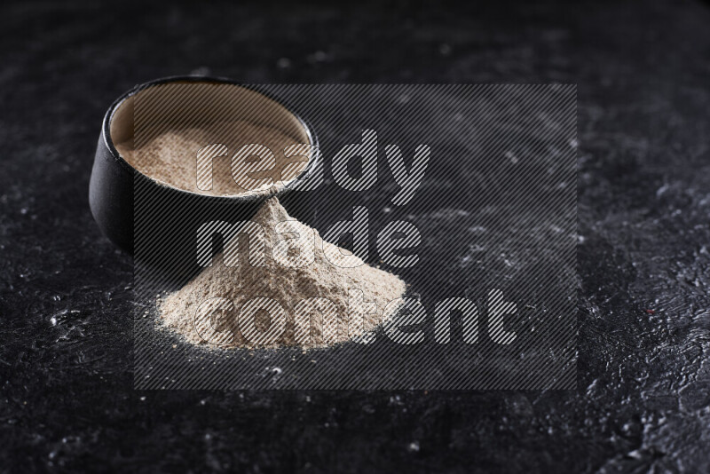 A black pottery bowl full of onion powder with fallen powder from it on black background