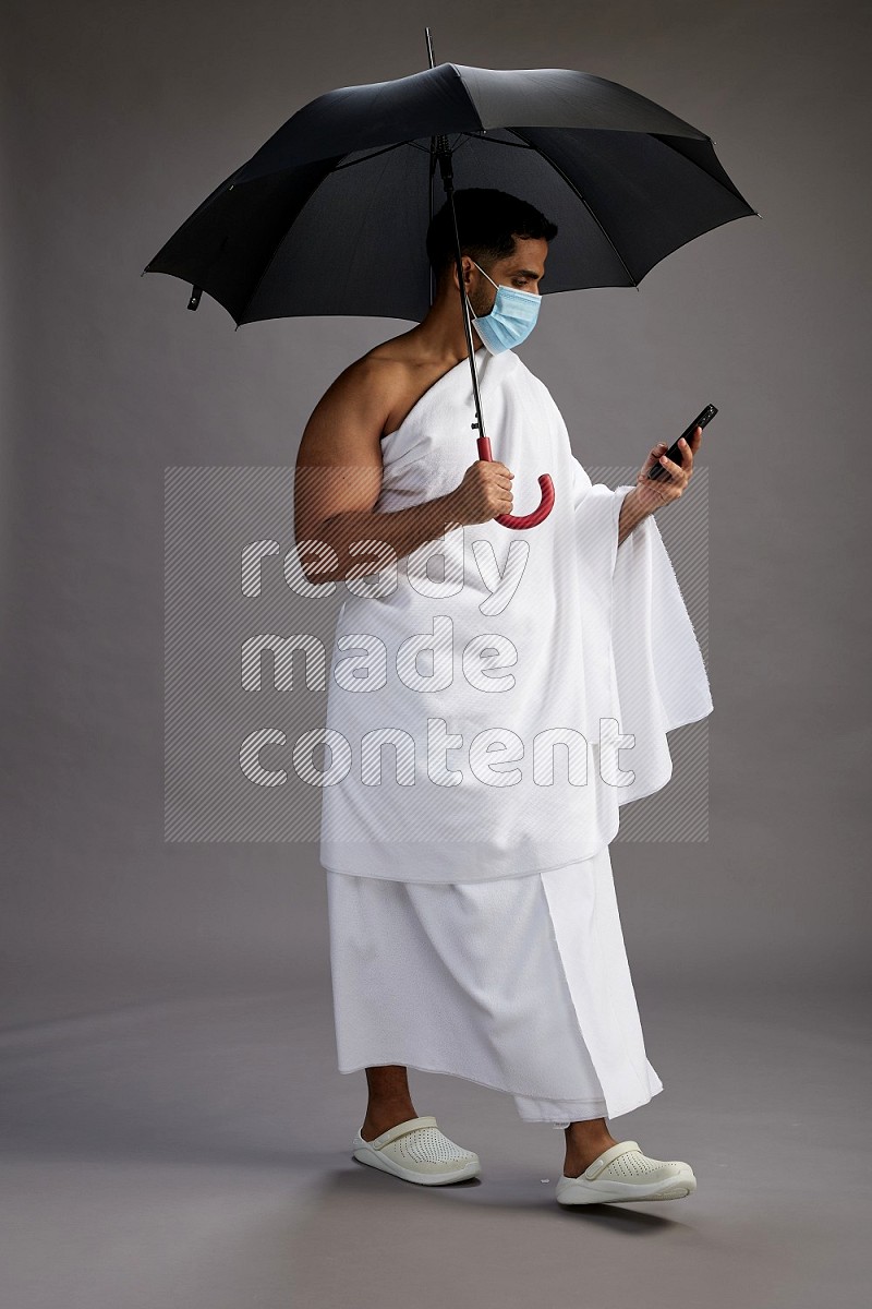 A man wearing Ehram and mask Standing holding umbrella on gray background on gray background