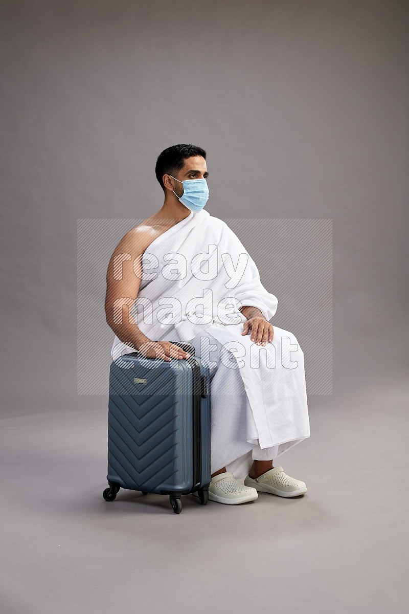 A man wearing Ehram sitting on chair holding traveling bag on gray background