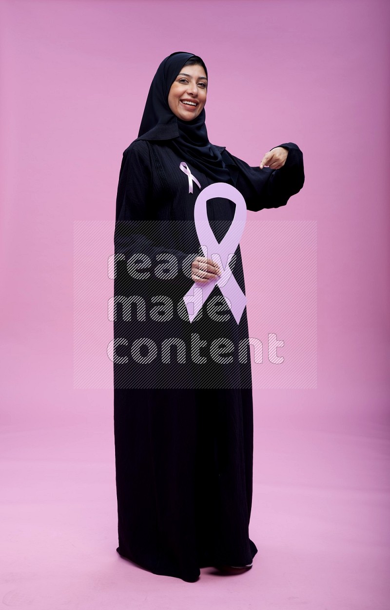 Saudi woman wearing pink ribbon on abaya standing holding awareness ribbon on plain pink background