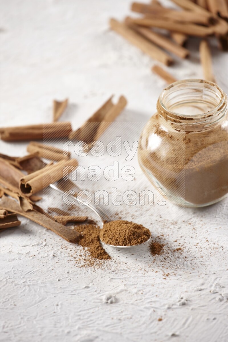 Herbal glass jar full cinnamon powder and a metal spoon surrounded by cinnamon sticks on a white background