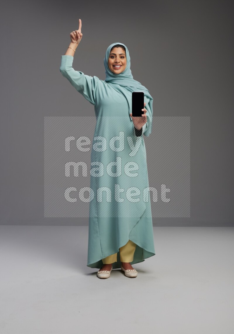 Saudi Woman wearing Abaya standing showing phone to camera on Gray background