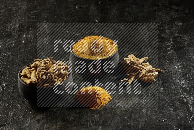 2 pottery black bowls, one full of turmeric powder and the other full of dried turmeric whole fingers on a textured black flooring