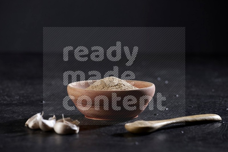 A wooden bowl and spoon full of garlic powder and beside it garlic cloves on a textured black flooring in different angles