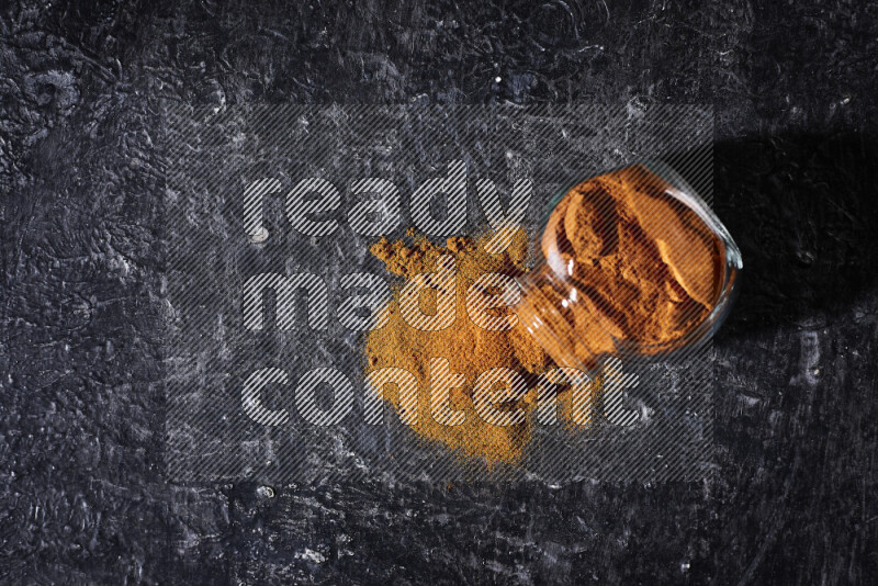 A glass jar full of ground paprika powder flipped with some spilling powder on black background