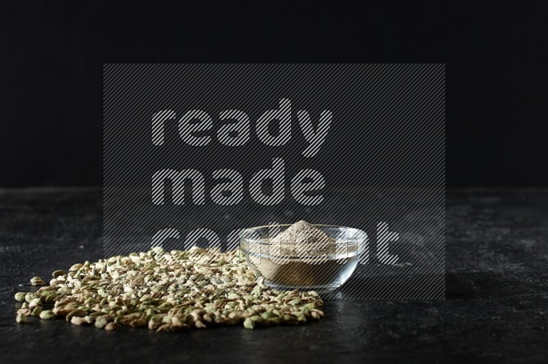 A glass bowl full of cardamom powder and cardamom seeds spreaded beneath the bowl on textured black flooring