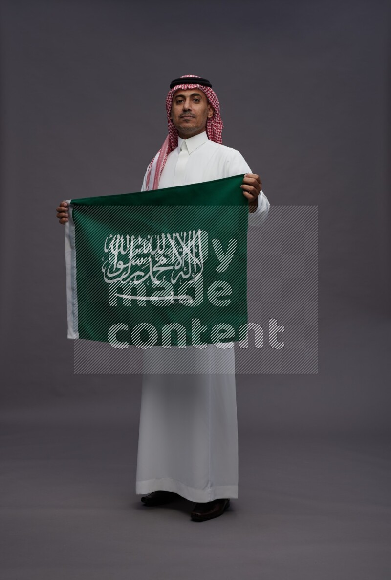 Saudi man wearing thob and shomag standing holding Saudi flag on gray background