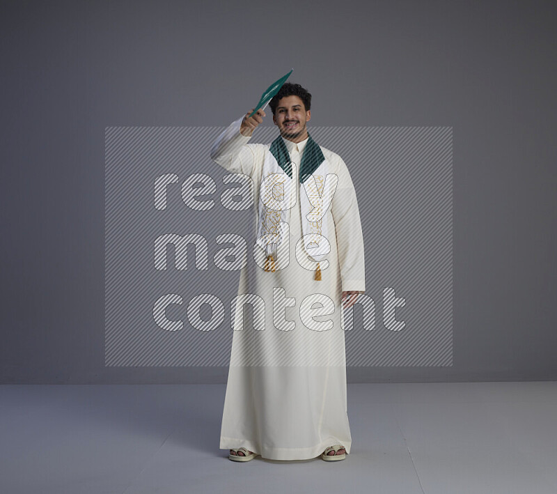 A Saudi man standing wearing thob and saudi flag scarf and holding small saudi flag on gray background
