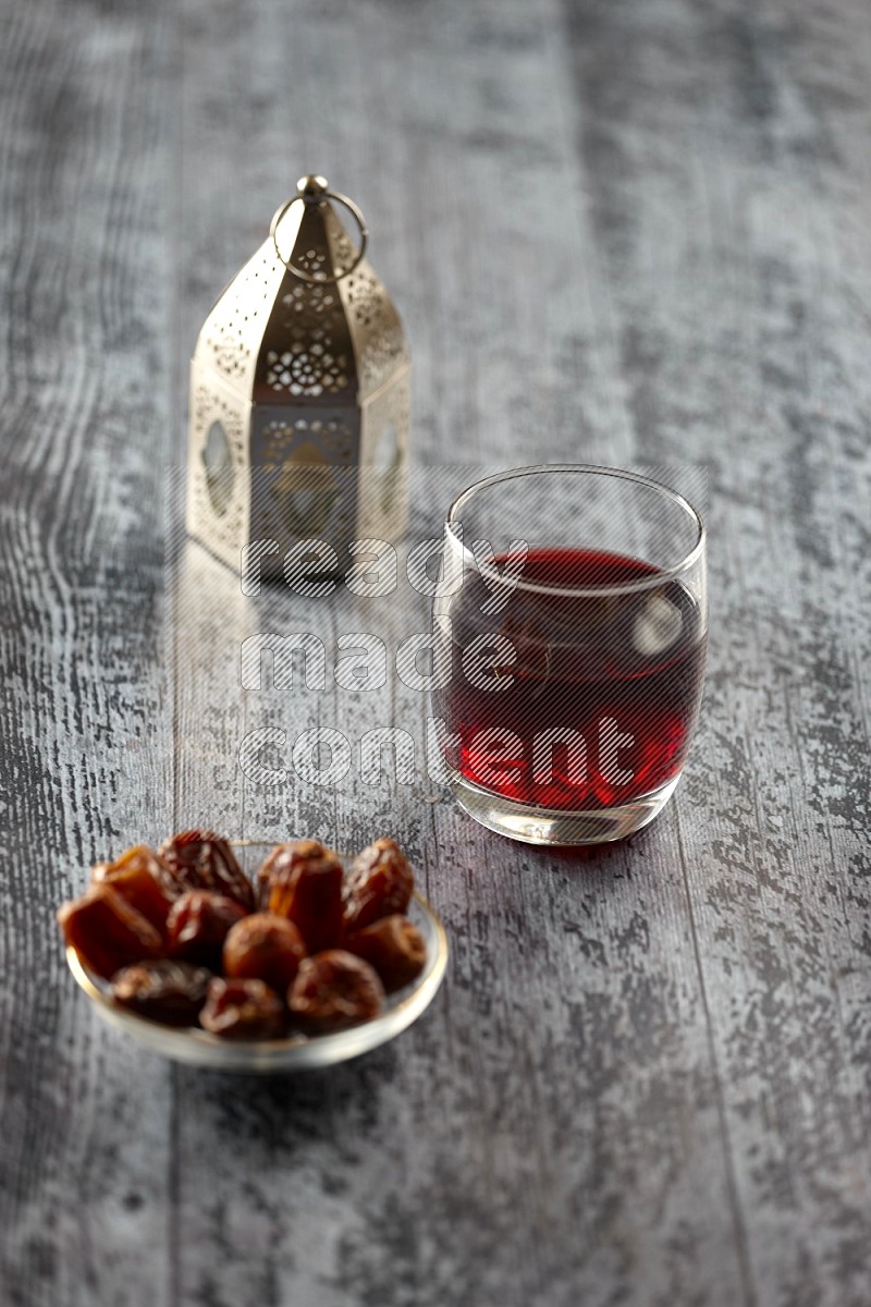 A silver lantern with different drinks, dates, nuts, prayer beads and quran on grey wooden background