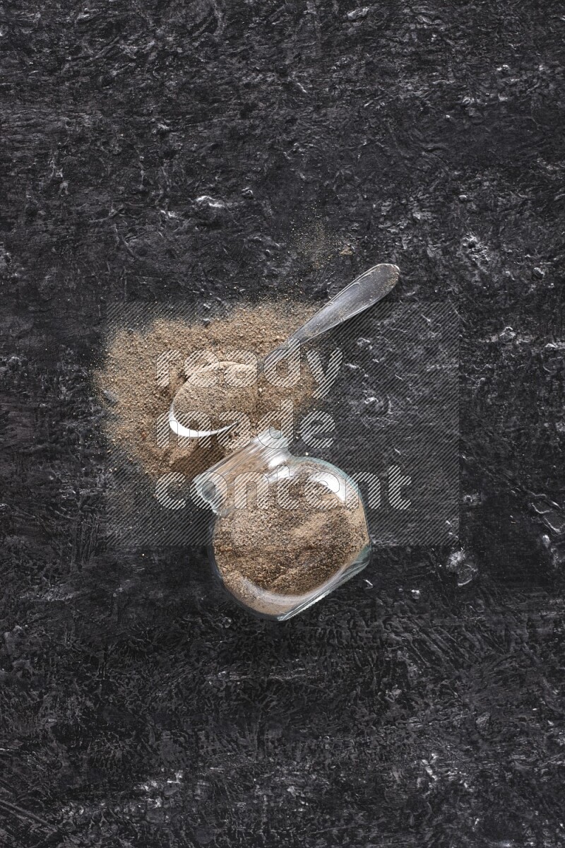 بودرة الفلفل الأسود في وعاء زجاجي مقلوب مع ملعقة معدنية مليئة بالبودرة علي خلفية سوداء