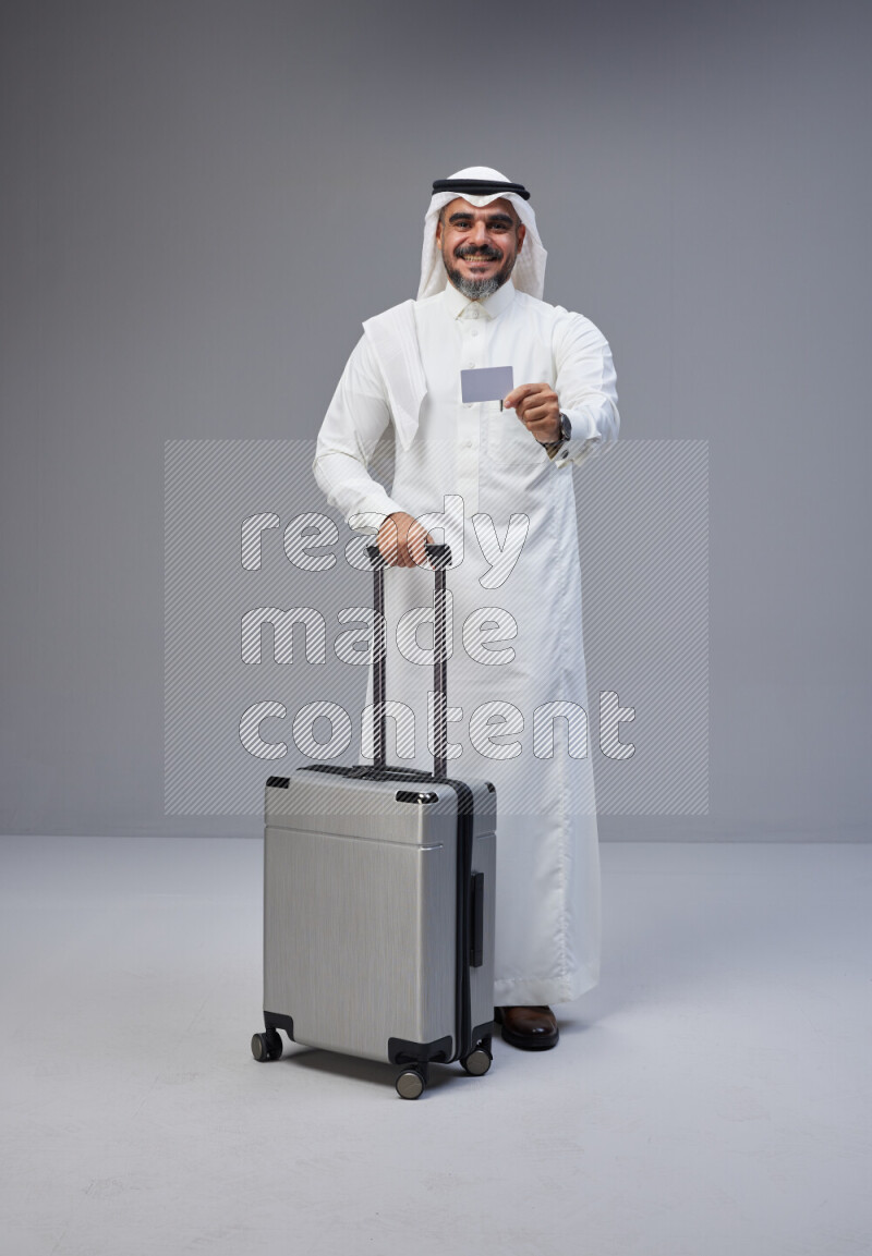 Saudi man wearing Thob and white Shomag standing holding Travel bag and ATM card on Gray background