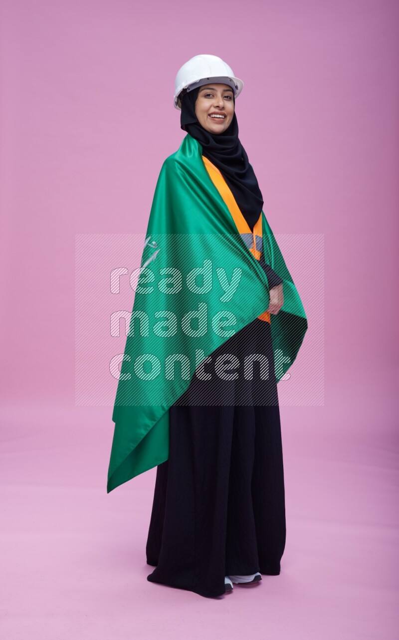 Saudi woman wearing Abaya with engineer vest and helmet standing holding Saudi flag on pink background