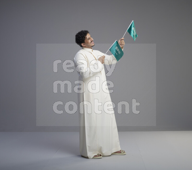 A Saudi man standing wearing thob holding small Saudi flag on gray background