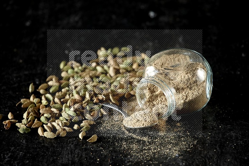 A flipped glass spice jar and a metal spoon full of cardamom powder and cardamom seeds spreaded on textured black flooring