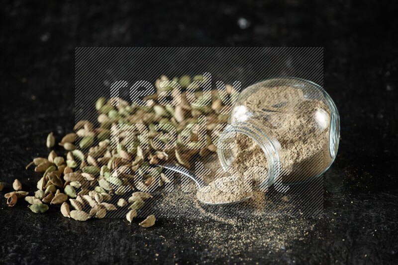 A flipped glass spice jar and a metal spoon full of cardamom powder and cardamom seeds spreaded on textured black flooring