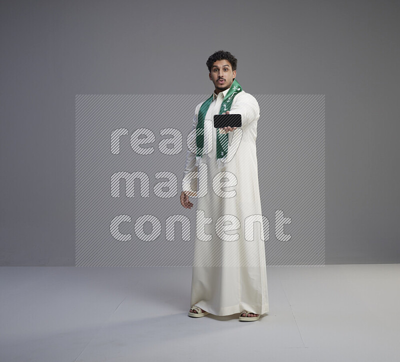 A Saudi man standing wearing thob and Saudi flag scarf showing phone to camera on gray background