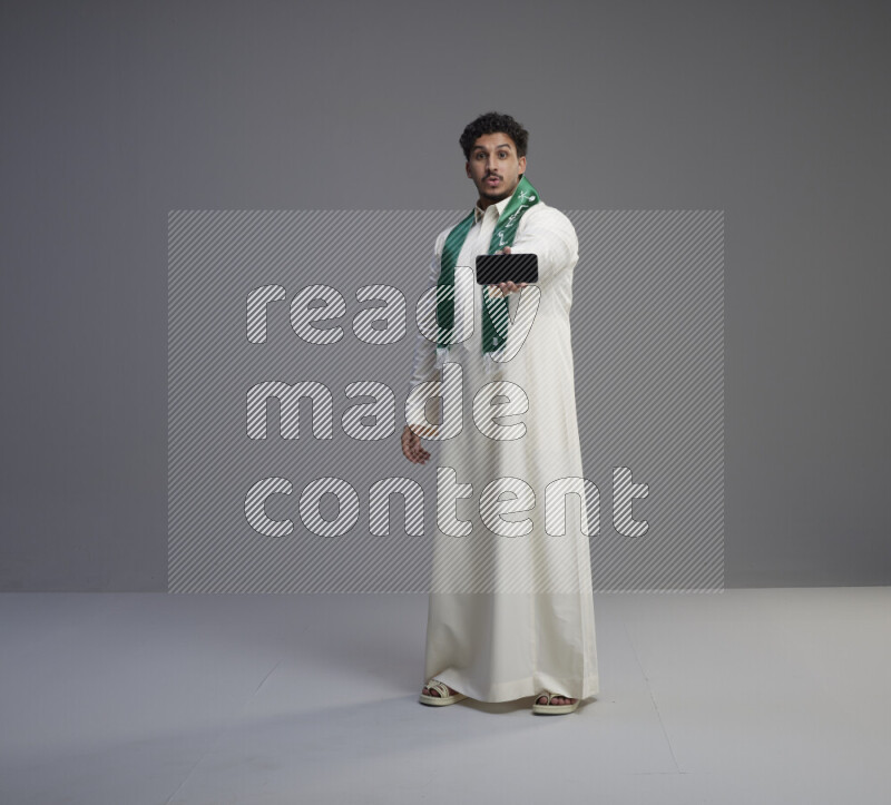 A Saudi man standing wearing thob and Saudi flag scarf showing phone to camera on gray background