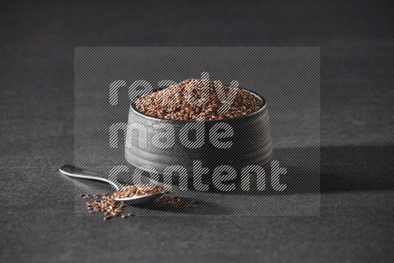وعاء فخاري أسود ممتلئ بحبوب بذر الكتان مع ملعقة معدنية ممتلئة على أرضية سوداء