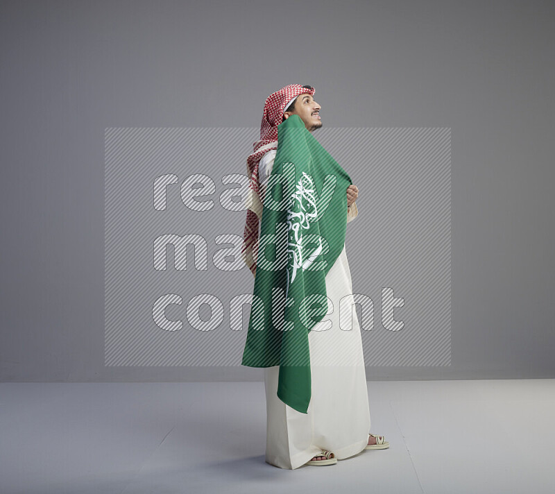 A saudi man standing wearing thob and red shomag holding big saudi flag on gray background