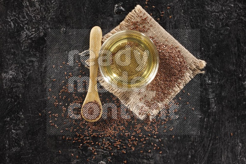 A glass bowl full of flax oil and wooden spoon full of flax with seeds spreaded on burlap fabric on a textured black flooring in different angles