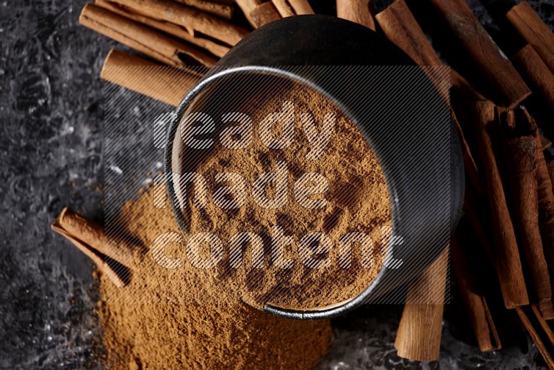 Black pottery bowl over filled with cinnamon powder and cinnamon sticks around the bowl on a textured black background