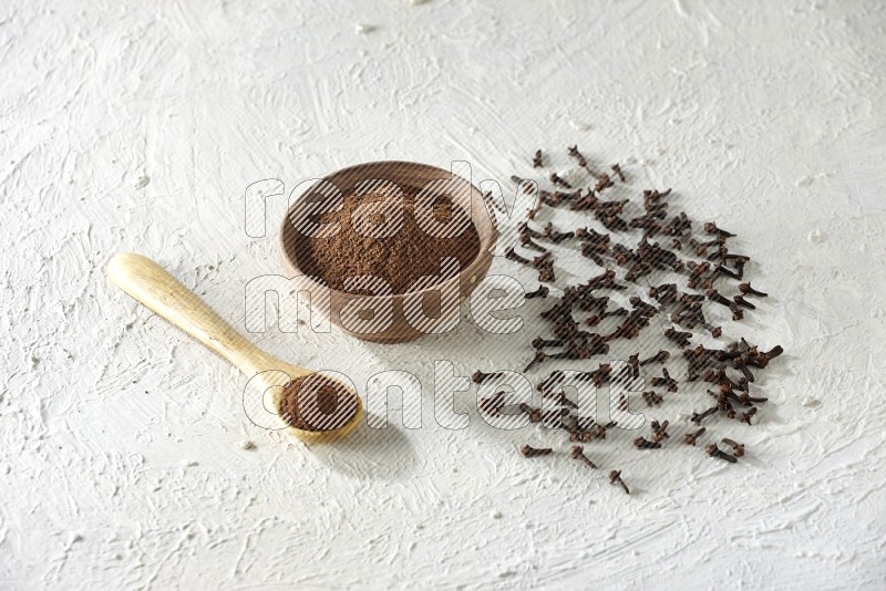 A wooden bowl and wooden spoon full of cloves powder with cloves spread on textured white flooring
