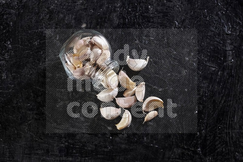 A glass spice jar full of garlic cloves flipped and the cloves came out on a textured black flooring in different angles