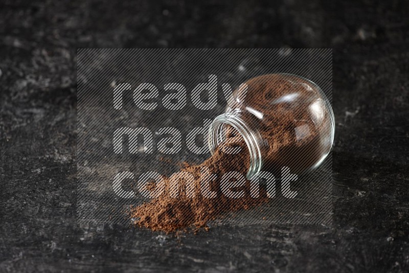 A flipped glass spice jar full of cloves powder and powder came out of it on textured black flooring