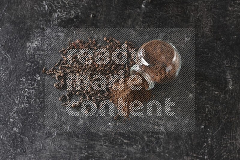 A flipped glass spice jar full of cloves powder and powder came out of it with cloves spread on textured black flooring