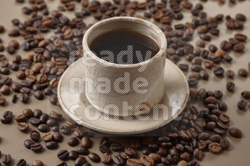 A beige pottery cup of coffee surrounded by roasted coffee beans on beige background
