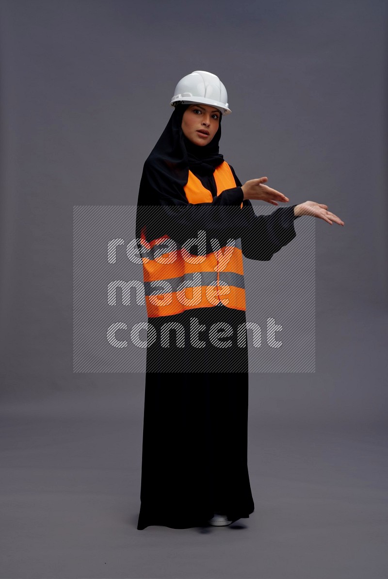 Saudi woman wearing Abaya with engineer vest standing interacting with the camera on gray background