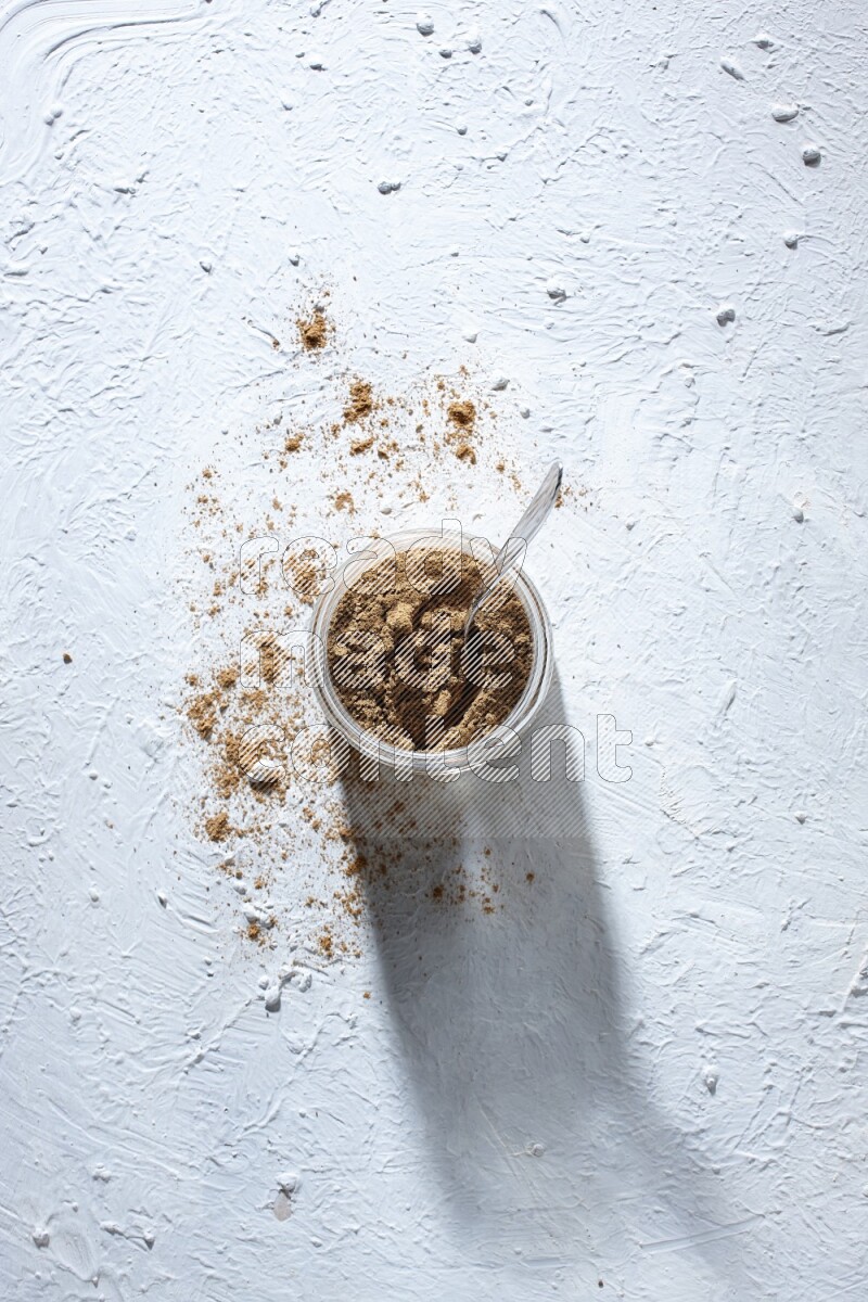 A glass jar and a metal spoon full of allspice powder on a textured white flooring