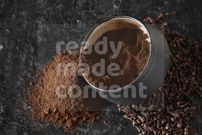 A black pottery bowl full of cloves powder spilled out of it with whole grains on a textured black flooring