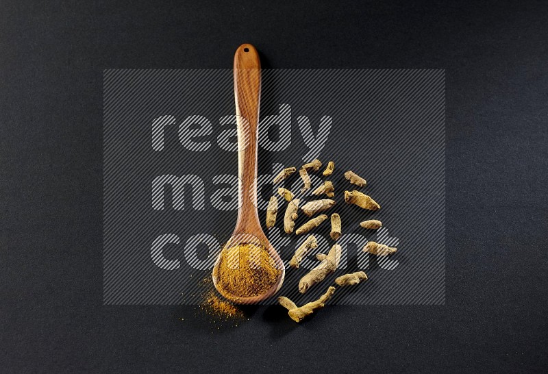 A wooden ladle full of turmeric powder with dried turmeric fingers on black flooring