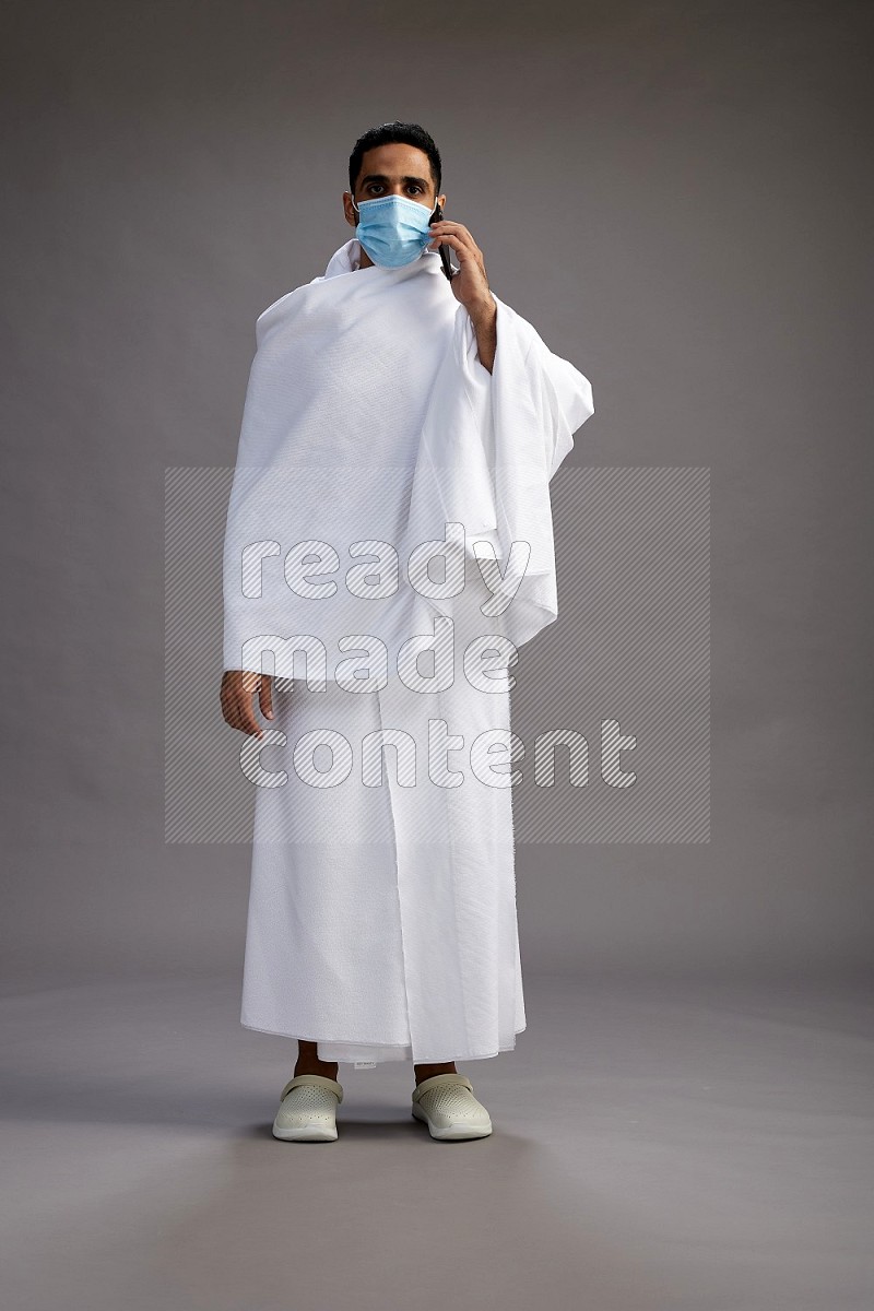 A man wearing Ehram with face mask Standing talking on phone on gray background