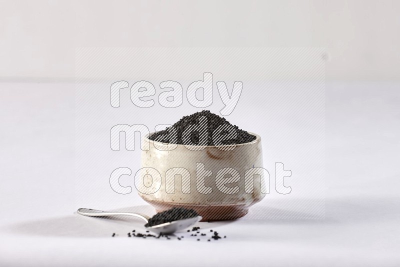 A beige pottery bowl and a metal spoon full of black seeds and more seeds spread on a white flooring