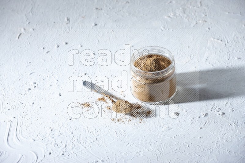 A glass jar and a metal spoon full of allspice powder on a textured white flooring