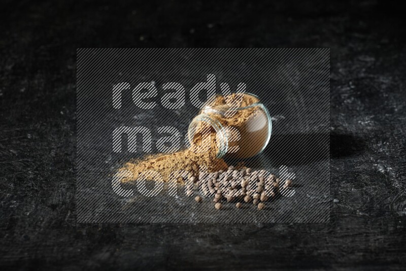 A flipped glass spice jar full of allspice powder and powder spilled out of it with whole balls on a textured black flooring